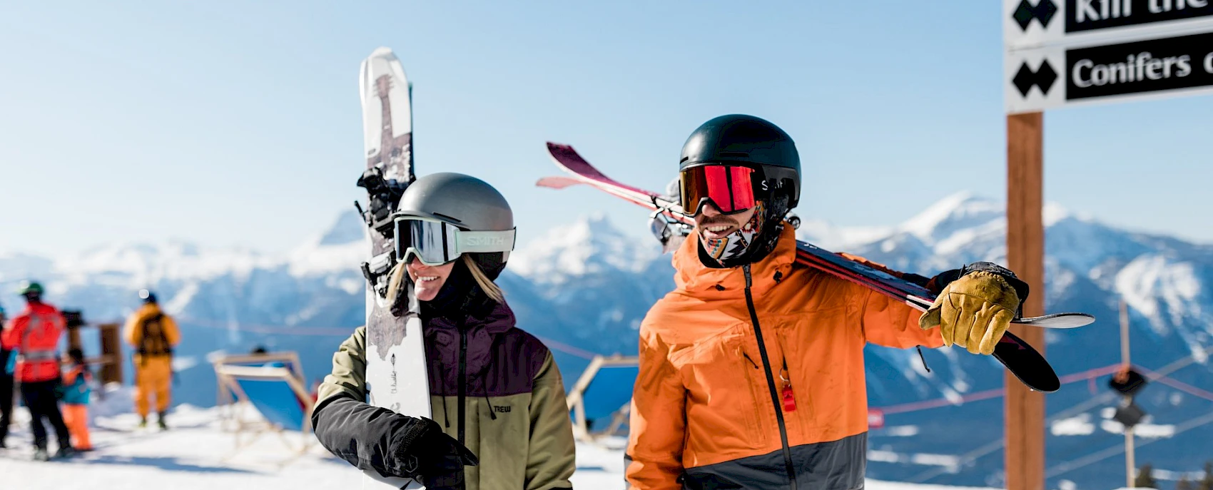 A man and woman smiling on a ski slope, holding skis, with mountains and a trail sign in the background