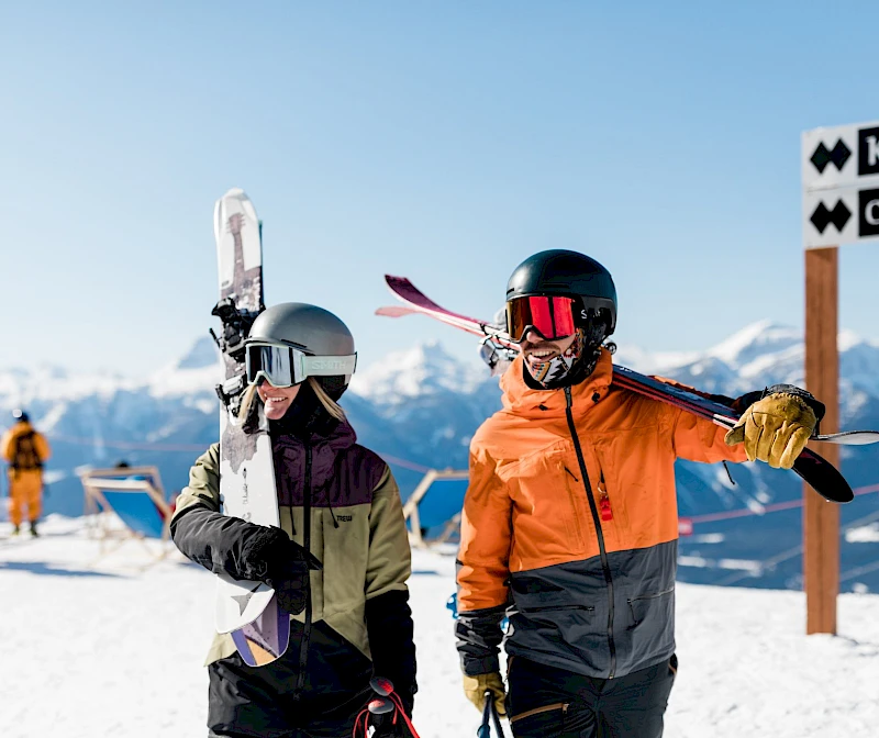 A man and woman smiling on a ski slope, holding skis, with mountains and a trail sign in the background